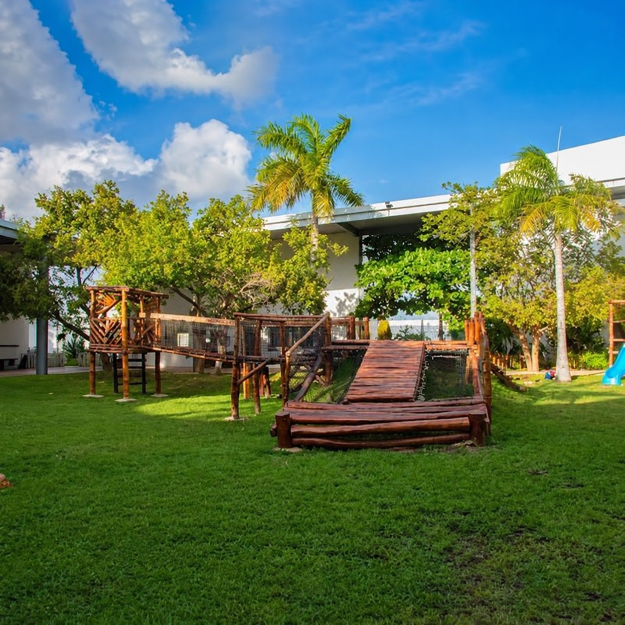 MACC ACADEMY — wooden playground and climbing structure set on a shaded green lawn beneath the Caribbean sky